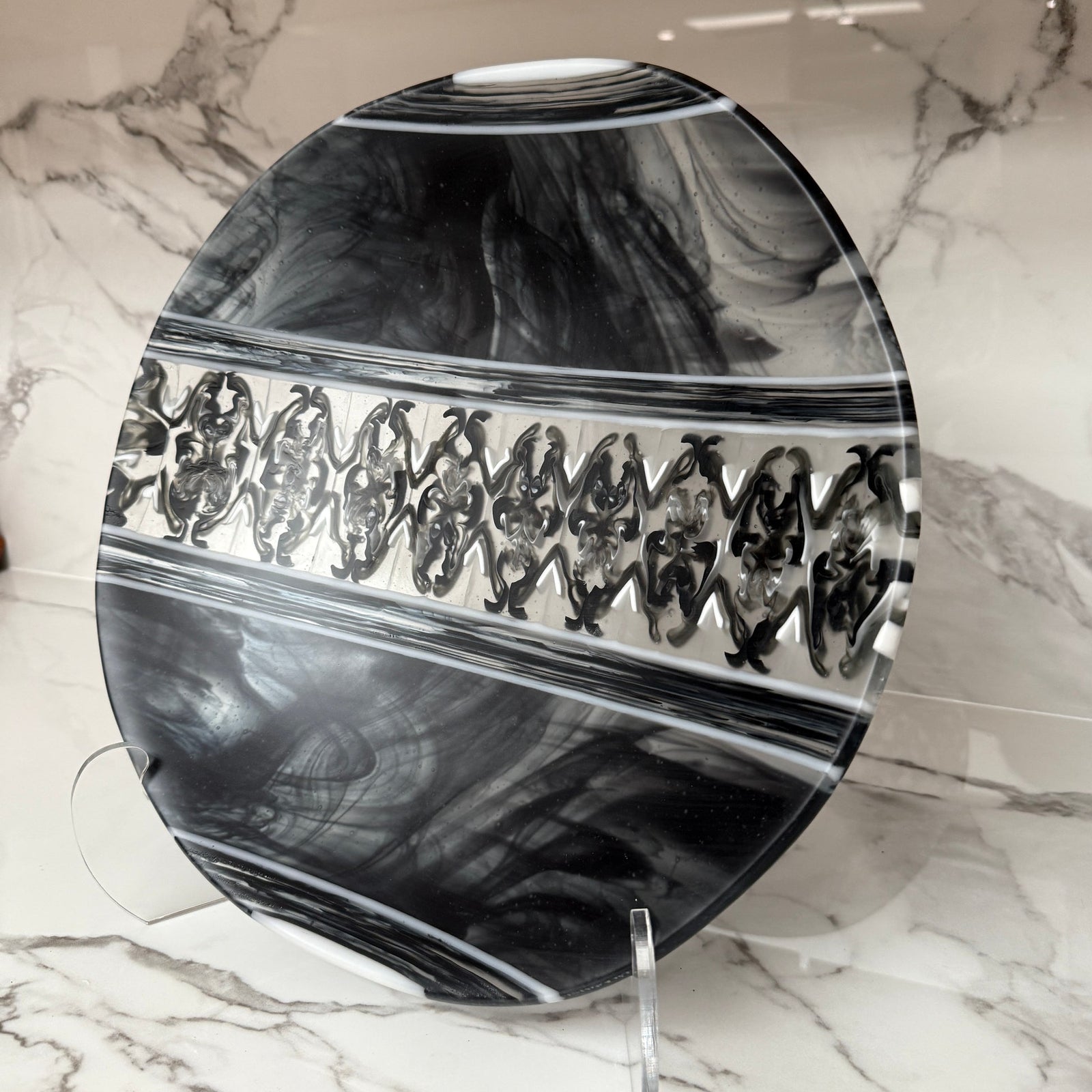 A round decorative plate with a glossy black and white marbled pattern and a central band of repeating black figures, displayed on a clear stand against a white marble-patterned wall.