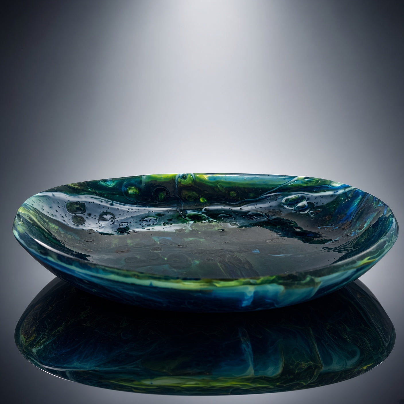 A shallow, wide glass bowl with a marbled pattern of deep blue, teal, and green, shown on a reflective surface against a dark gray gradient background.