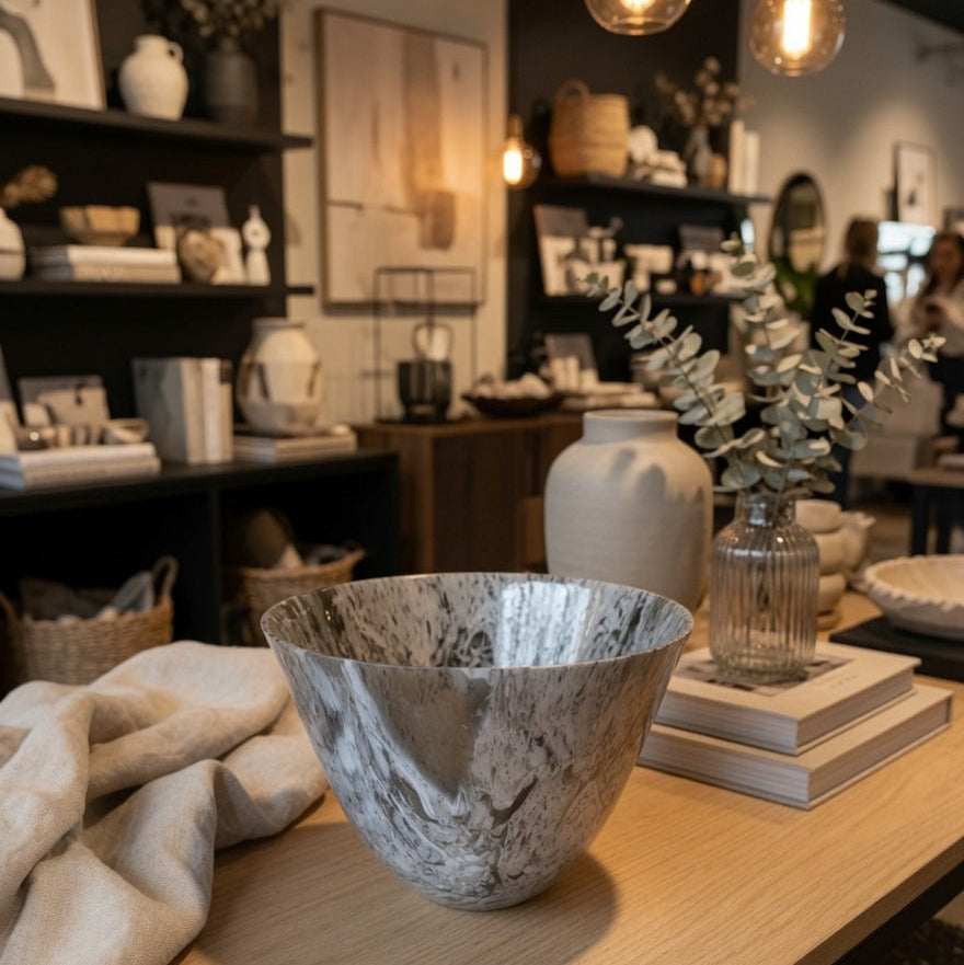A large round marble-look bowl with a gray and white veined pattern sits on a light wood table in a styled home decor store. A beige linen cloth, stacked books, a small glass vase with eucalyptus, and other decor items are arranged around it, with warm hanging bulbs and shelves visible in the background.