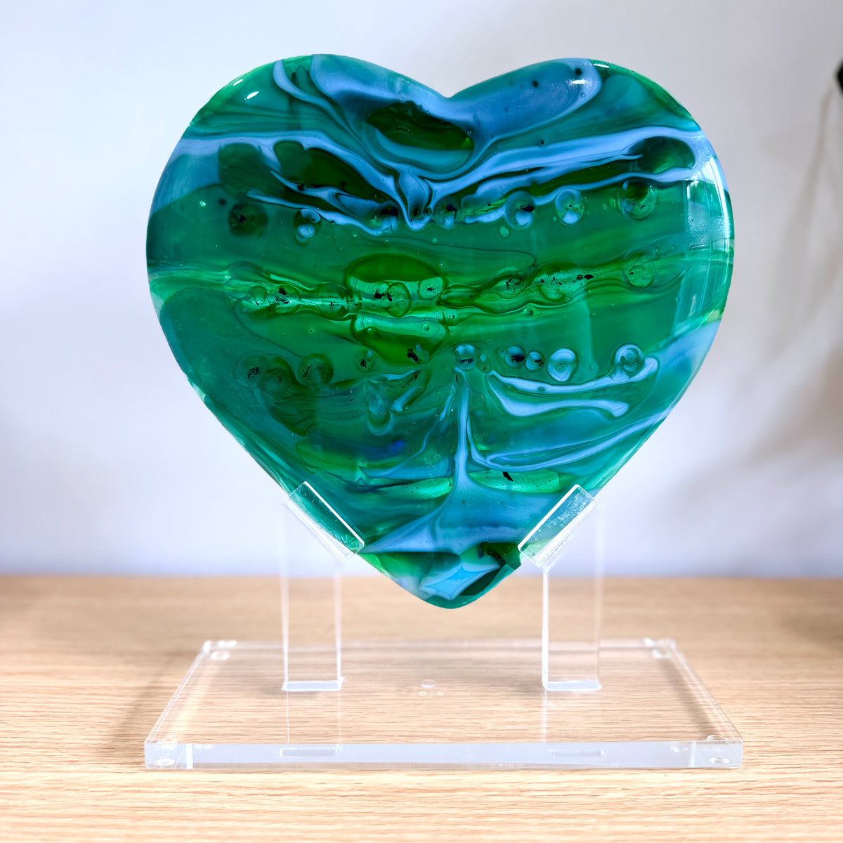 Emerald Tide green and blue fused glass heart sculpture on a clear acrylic stand on a wood table - handmade art glass sculpture