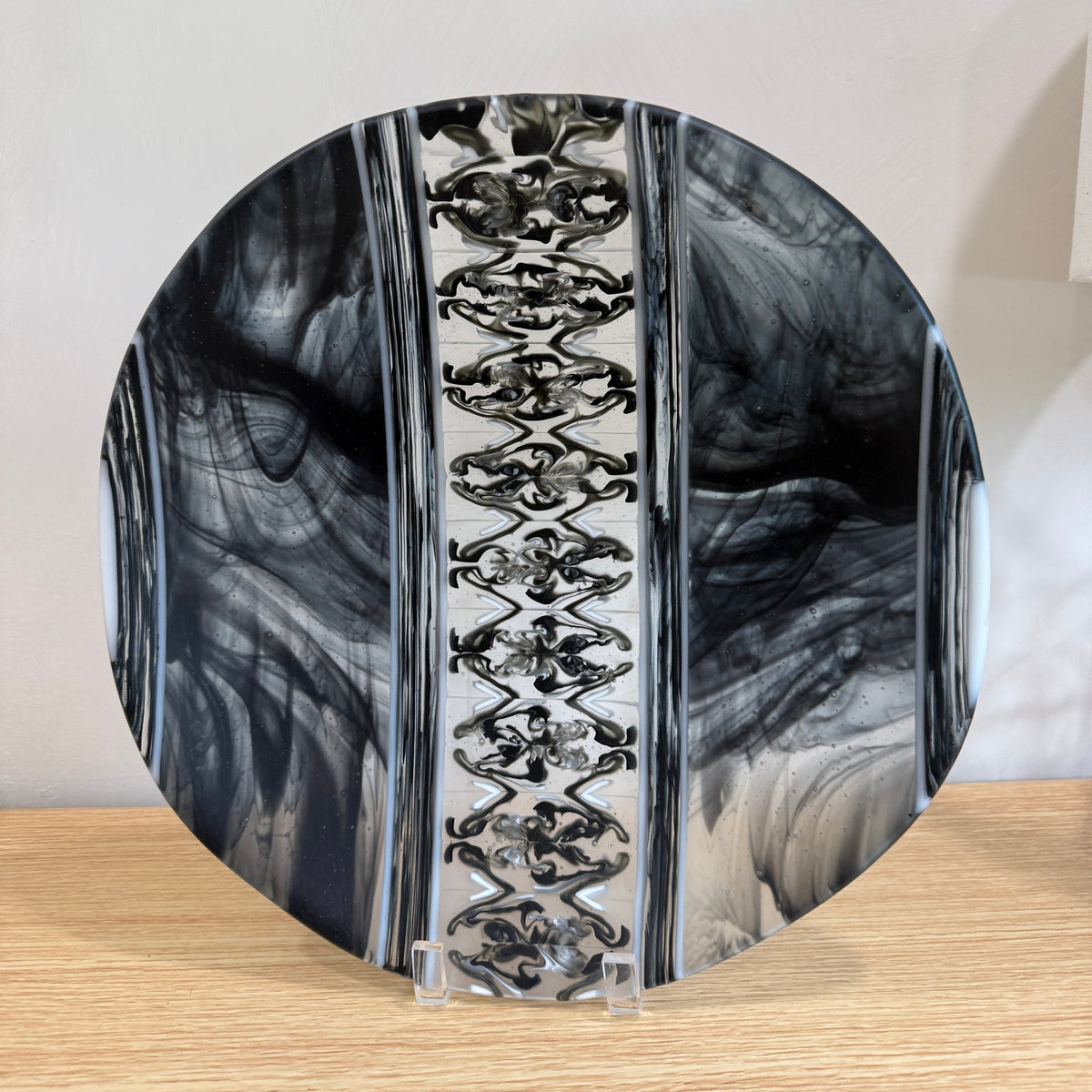 A round decorative plate with a black, gray, and white marbled pattern and a central vertical band of repeating silver filigree, displayed on a clear stand on a light wood surface against a white wall.