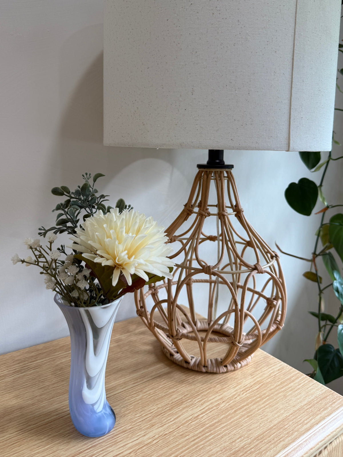 Lamp with wicker base and white shade on a wooden surface with a vase and flowers.
