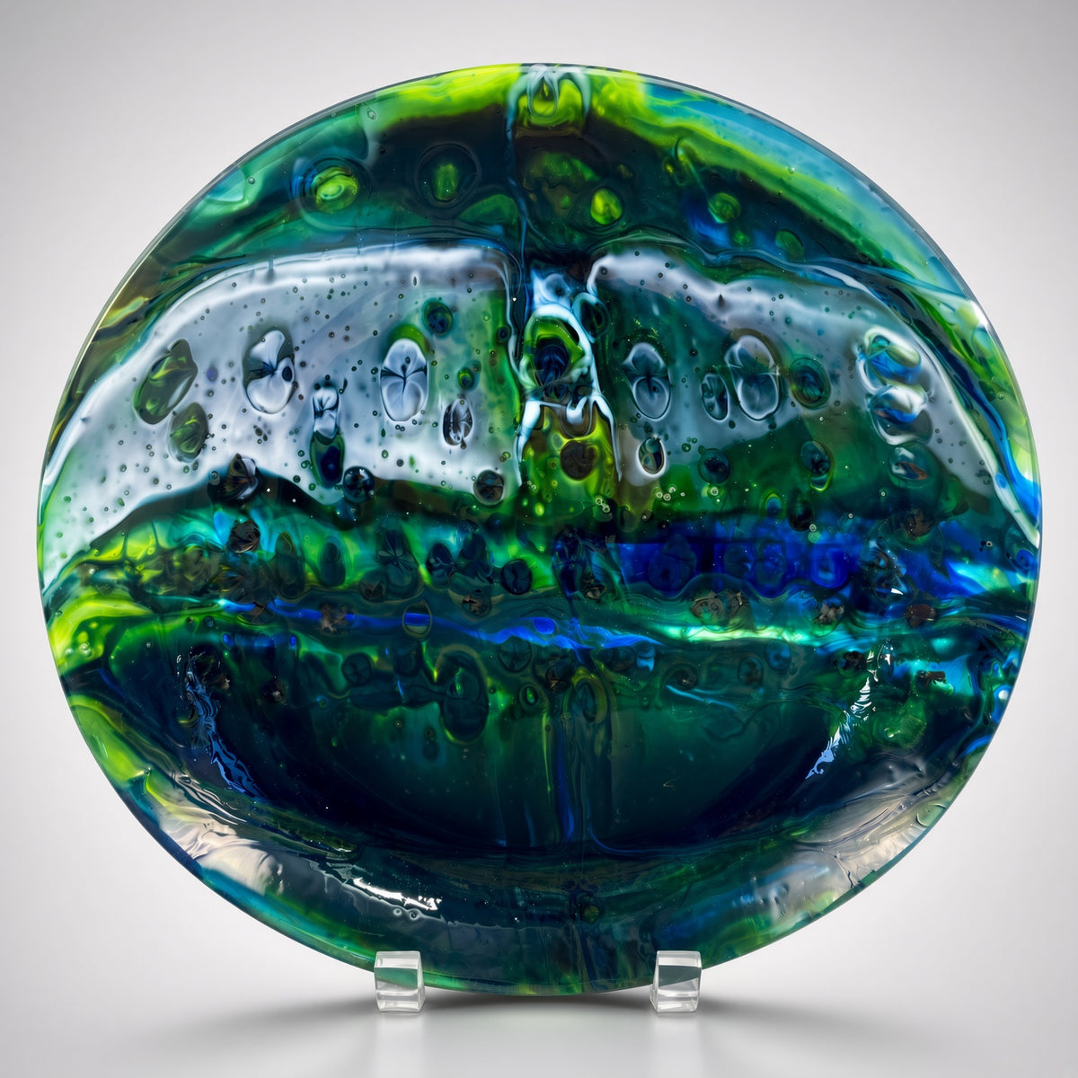 A round glass plate with a highly textured, organic pattern of green, blue, and white, displayed on a clear acrylic stand against a light gray background.