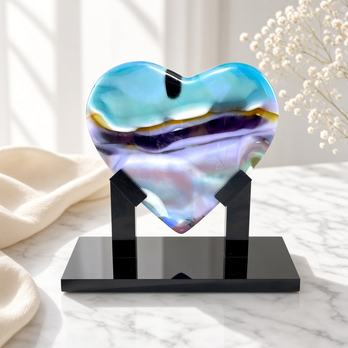 Aqua, purple, and amber fused glass heart sculpture on black stand, staged on a marble table with pampas grass - Arctic Skies handmade art glass gift