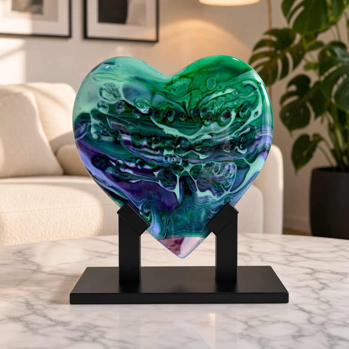 A heart-shaped glass sculpture with swirling green, teal, and purple patterns, displayed on a black metal stand on a marble table in a modern living room setting.