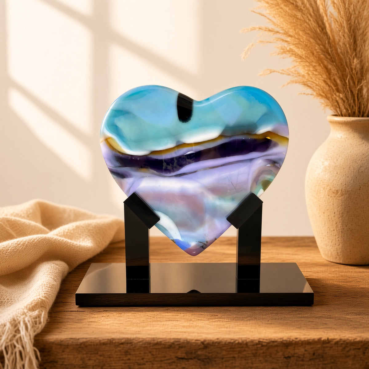 Aqua, purple, and amber fused glass heart sculpture on black stand, staged on a wooden table with vase and cream blanket - Arctic Skies handmade art glass gift