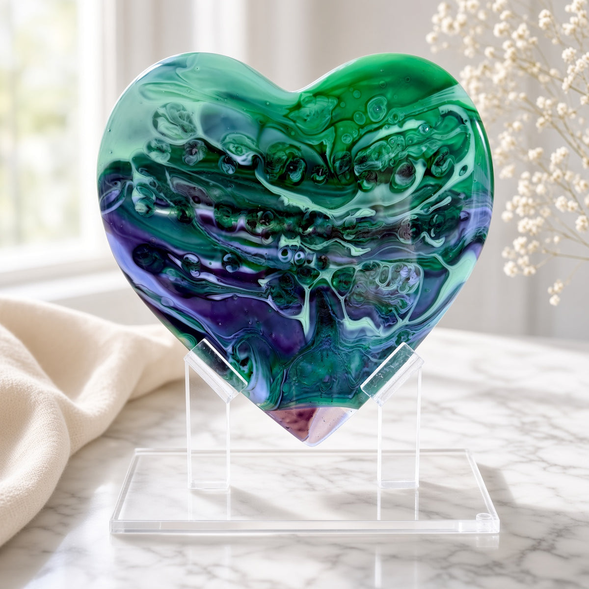 Green, purple, and blue marbled glass heart sculpture on a clear acrylic stand on a marble surface - handmade fused art glass keepsake