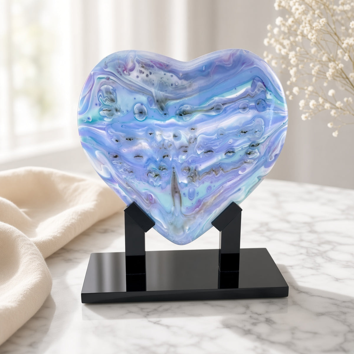 Pastel lavender, sky blue, and teal fused glass heart sculpture on black stand, staged on a marble table with pampas grass - handmade art glass gift
