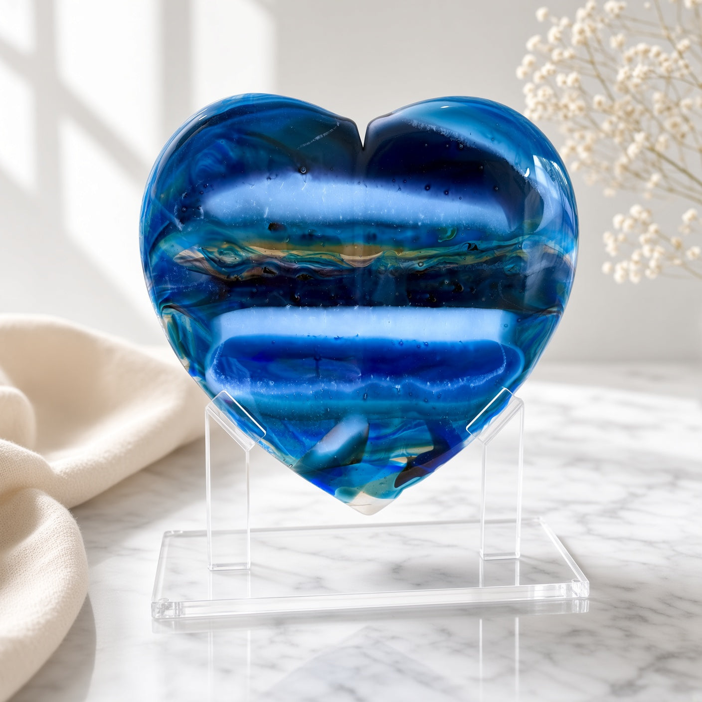 XL blue banded fused glass heart sculpture on a clear acrylic stand, styled on a marble surface with soft linen and dried flowers — handmade art glass decor.