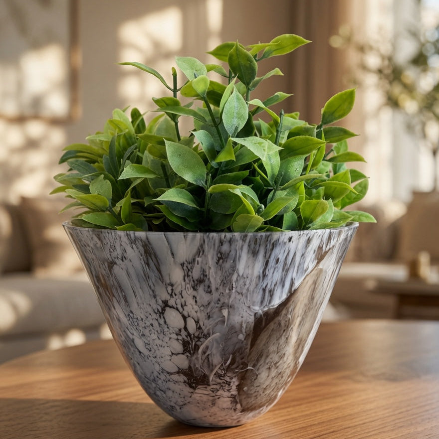 A green leafy plant in a wide, sculptural ceramic pot with a gray and white marble pattern, displayed on a wooden table in a warm, softly lit living room.